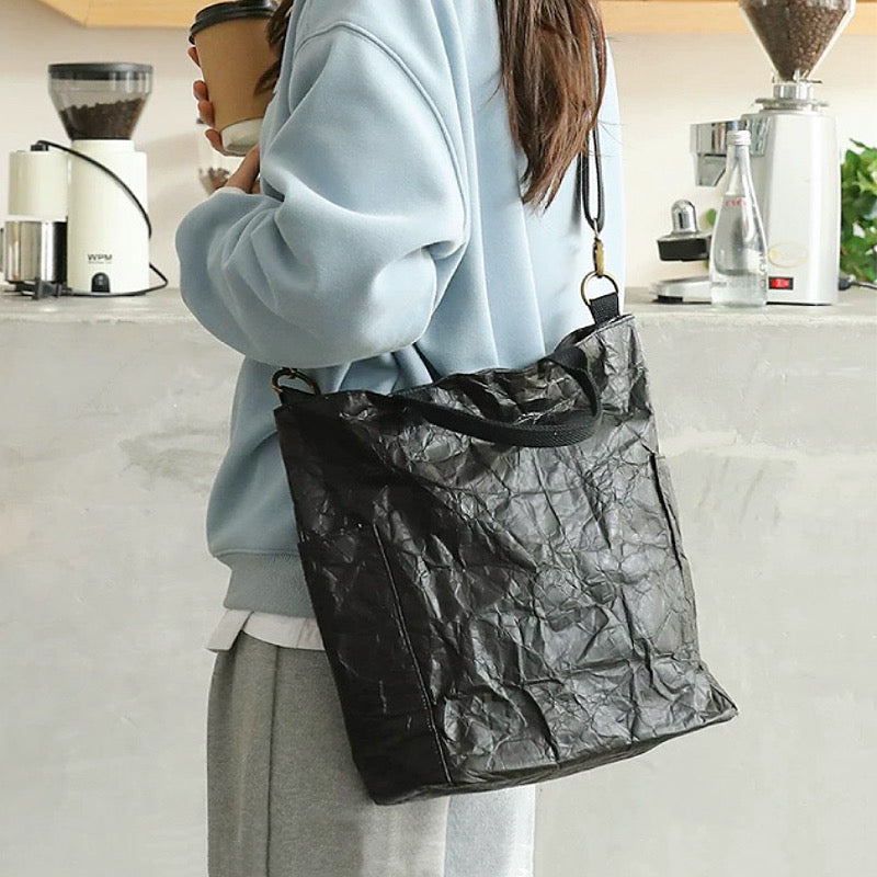 A model wearing a black kraft-paper-textured tote bag as crossbody, showcasing a modern urban explorer look.