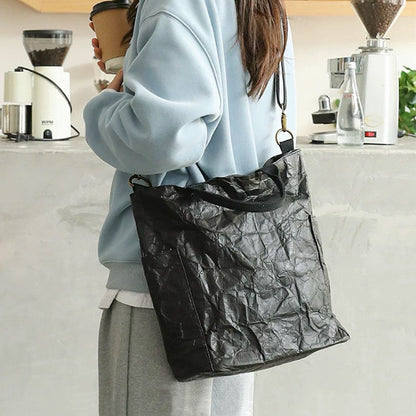 A model wearing a black kraft-paper-textured tote bag as crossbody, showcasing a modern urban explorer look.
