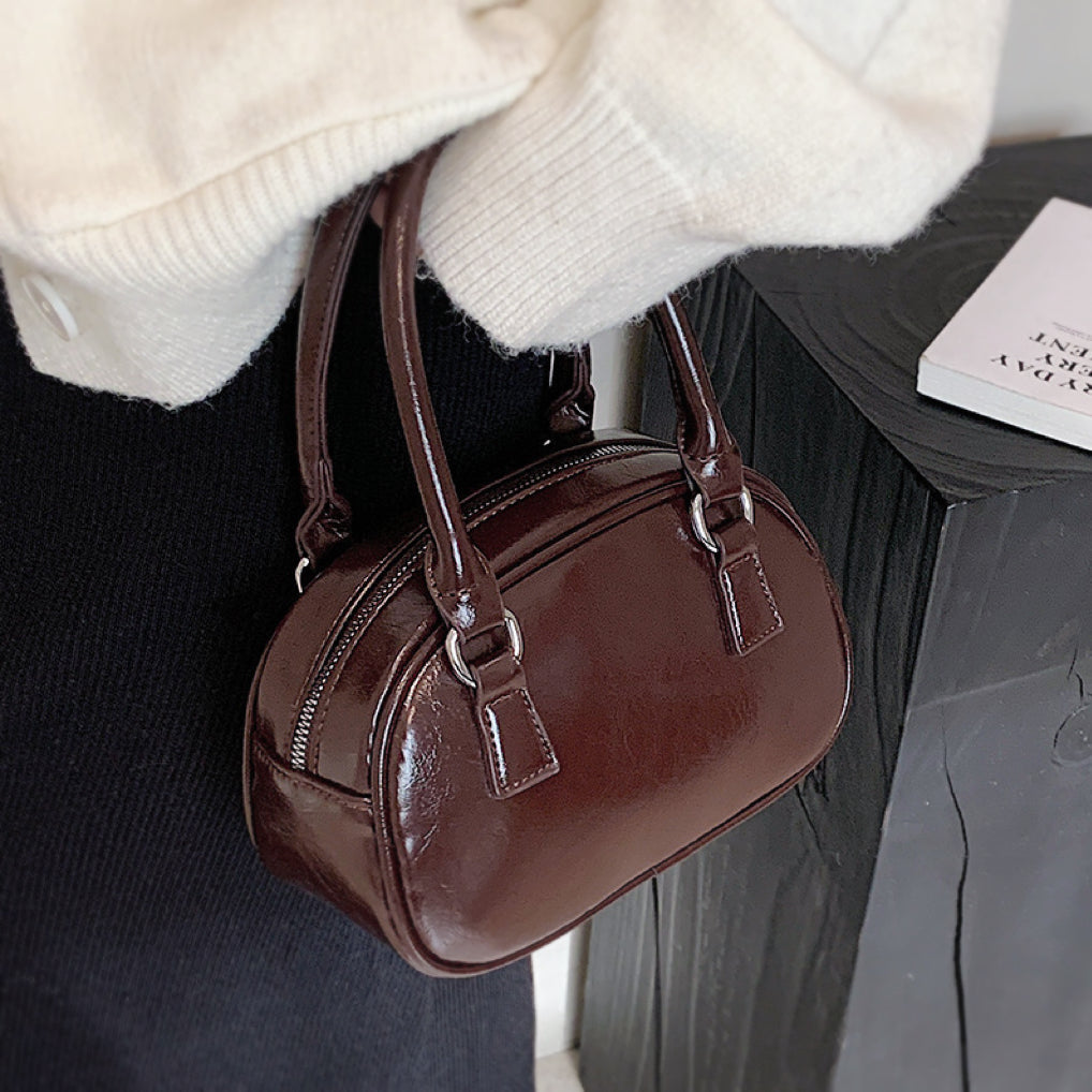Angled view of umber PU leather mini bowling bag with grab handle and zip-top fastening .