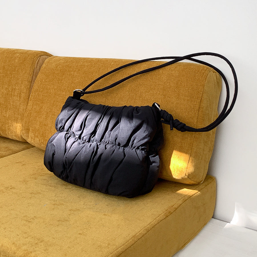 Front view of Black pleated  cloud bag with an adjustable strap, resting on a yellow couch Pleated Cloud.