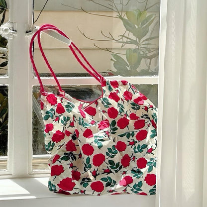 Angled view of Polyester Rose Romance Tote Bag with red rose print, red grab straps, hanging in a window.