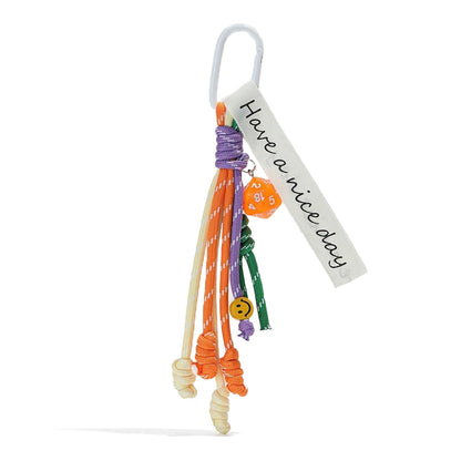 Front view of Polyester tassel bag charm with white carabiner, Have a nice day tag, orange dice, and smiley face.